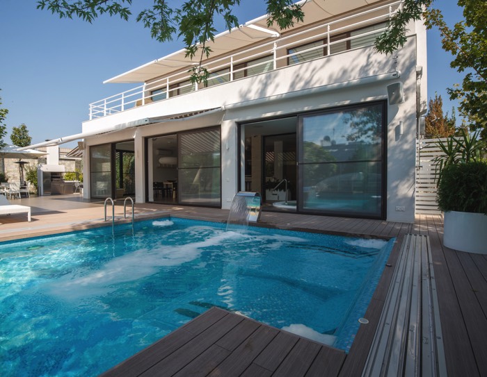 Aménagement d'une piscine moderne avec petit escalier et fontaine. Parquet tout autour avec habitation blanche moderne et grandes baies vitrées.