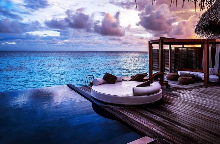 Aménager une piscine en bois wengé juste au bord de la mer. Salon de jardin avec canapé rond blanc et plusieurs poufs à proximité sous petite tonnelle.