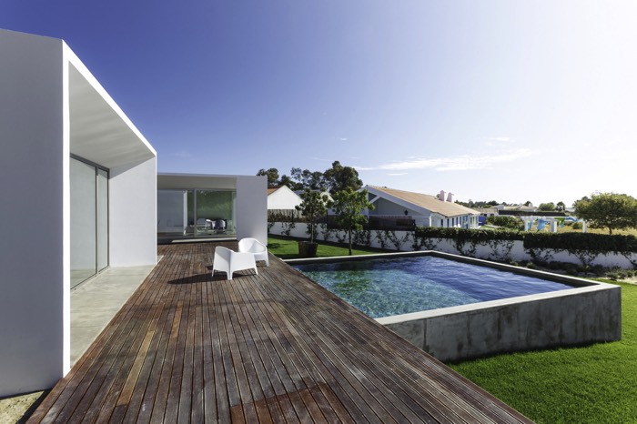 Aménager une piscine carré en béton collé à une terrasse en parquet avec deux petits fauteuils dessus. Maison au style architectural moderne original blanc avec baies vitrées et pelouse.