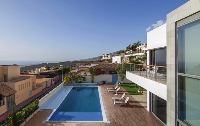 Aménagement d'une piscine rectangulaire en bois chêne avec dalles blanches et grises tout autour. Quatre transats et maison moderne blanche.