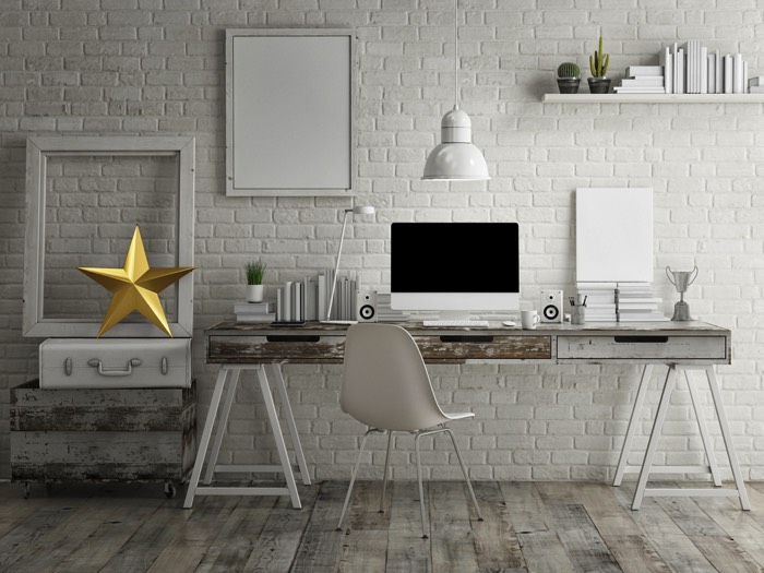 Aménager un bureau en bois avec tiroirs dans une pièce moderne blanche avec un mur en pierre. PC, petites baffes, tableau blanc et étagère en haut à droite.