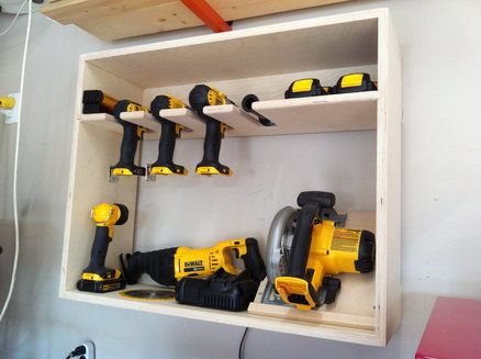 Rangement rectangulaire en bois utile et pratique pour ranger une multitude de perceuses jaune et noires ainsi qu'une tronçonneuse. Mur blanc uni.