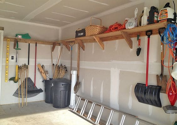 Rangement bois en hauteur utile en L avec multitude d'outils accrochés comme une pelle et un mètre. Pièce avec peinture en cours, poubelles noires au fond.