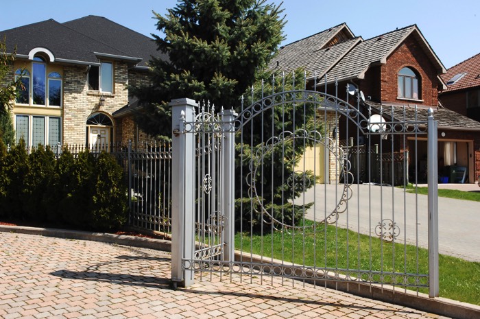 Portail aluminium contemporain blanc avec petits barreaux verticaux et pointes en hauteurs. Pelouse, sapin et allée goudronnée donnant vers deux maisons.