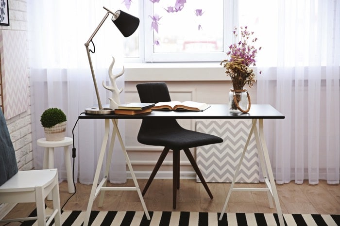 Aménagement d'un petit bureau classique élégant avec livres et fleurs dessus. Fauteuil noir, pièce avec parquet, tapis noir et blanc ainsi que rideaux transparents.