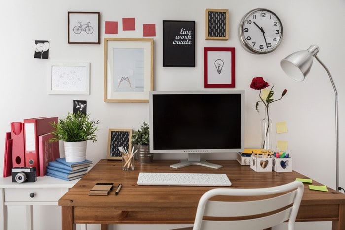 Aménager un bureau en bois noyer avec PC, rose rouge et clavier blanc posé dessus. Multitude de tableaux différents, horloge, lampe et table blanche.