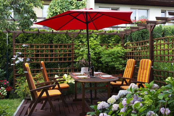 Aménager une terrasse avec table ovale avec quatre chaises oranges et parasol rouge. Clôture en bois, pelouse et végétation abondante style green power. Aménagement d'une terrasse2