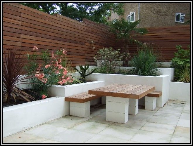 Parpaings décoratifs blancs servant de piliers pour une table d'extérieur en bois et ses bancs. Clôture en bois, végétation et palmiers.
