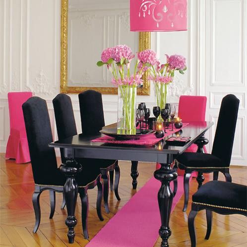 Salle à manger blanche composée d'une table et de chaises noires ajustées par les accessoires de couleur fushia. Parquet et grand miroir.