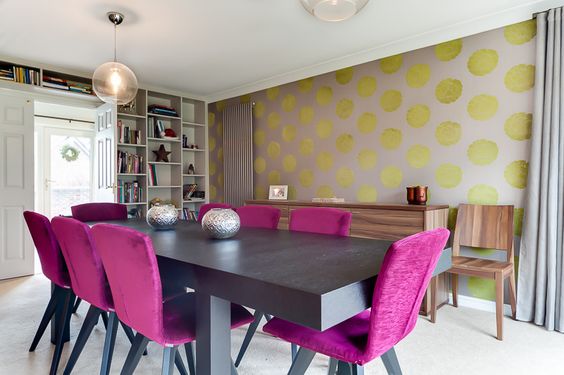 Salle à manger à l'ambiance pop composée d'une table en bois et de chaises couleur fushia. Mur avec ronds jaunes or sur fond beige.