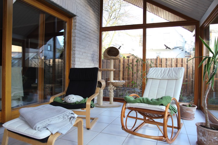 Véranda moderne en verre et en bois sapin avec carrelage blanc classique. Chaise à bascule blanche, chaise noire et baie vitrée en verre.