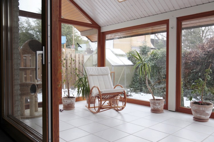 Véranda moderne blanche en bois sapin et en verre avec carrelage et vue sur le jardin. Fauteuil à bascule blanc avec plusieurs plantes.