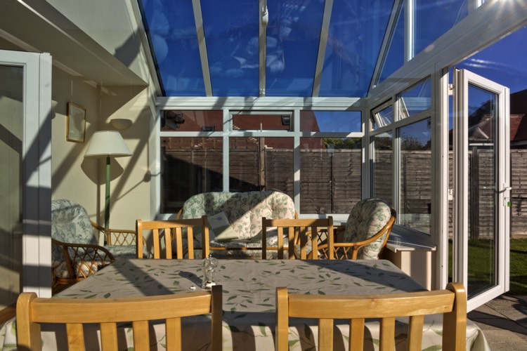 Véranda moderne blanche en verre avec table à manger rectangulaire et quatre chaises en bois bouleau. Petit salon avec canapé et fauteuils.