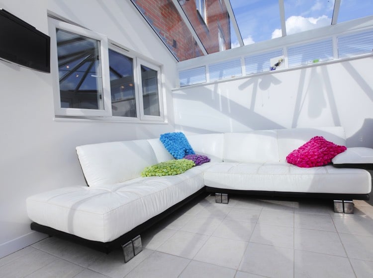 Véranda moderne blanche style pop avec canapé en L et quatre coussins de couleurs différentes. Carrelage blanc classique et fenêtre.