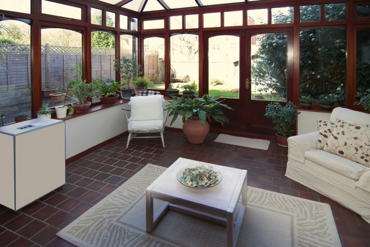 Véranda moderne en verre et en bois noyer avec vieux carrelage. Plan de travail en L, petit salon avec table carré sur tapis moderne et fauteuil blanc.