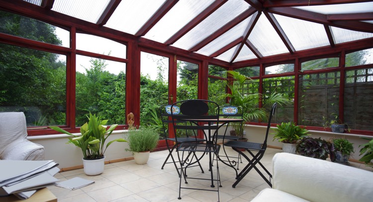Véranda moderne en verre de couleur rouge foncé avec carrelage. Petite table ronde avec chaises vertes foncés en acier ainsi qu'une multitude de plantes donnant un côté green power.