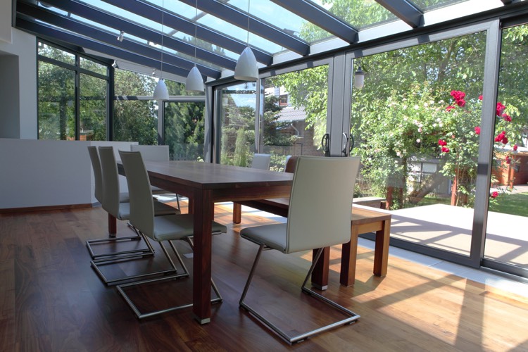 Véranda moderne blanche en verre avec parquet stratifié et vue sur le jardin avec rosier. Table rectangulaire en bois avec chaises blanches modernes et minimalistes.