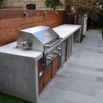 Cuisine extérieure béton avec barbecue intégré à côté du plan de travail. Rangements en bois noyer et wengé sur carrelage blanc et gris.