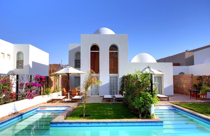 Aménagement d'une piscine extérieure en U avec petit jardin composé d'arbustes et de rosiers. Parasols, transats avec maison blanche style marocain.