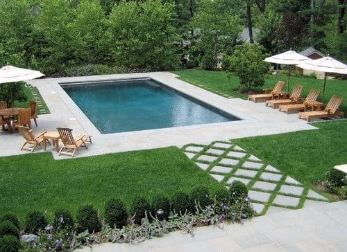 Aménagement d'une piscine avec jardin composé d'arbre et d'une pelouse. Allée en pierres en forme de losange, transats en bois et parasols blancs.