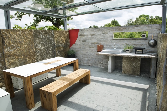 Aménagement d'une cuisine d'été ouverte avec pergola en verre et en pierre. Petite table en bois rectangulaire avec banc et plan de travail rustique en pierre.