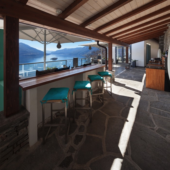 Aménagement cuisine ouverte avec plan de travail en bois. Pergola, bar en bois avec quatre sièges de bar en bleu clair et panorama sur la mer.