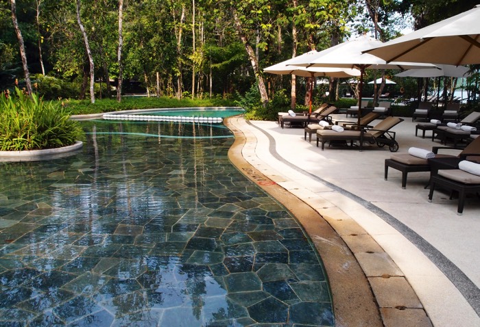 Jardin aménagé autour d'une piscine dont le sol est en pierre. Des transats surmontés de grands parasols sont étendus d'un côté de la piscine.