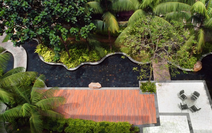 Photo prise en hauteur d'un jardin aménagé composé d'arbres et d'un palmier. Il comprend également une rivière et une terrasse en pierre avec une table ronde entourée de quatre fauteuils.