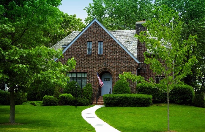 Maison en briques du style colonial au moderne doté d'une baie vitrée, d'une porte blanche et d'une cheminée. Petite allée sur pelouse verte et tondue avec arbres.