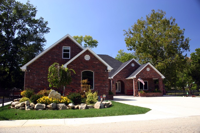 Maison en briques rouges style colonial avec plusieurs fenêtres et porte blanche. Clôture noire, végétation, plantes et multitude d'arbres.