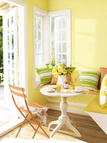 Salle à manger jaune clair avec parquet en érable. Petite table ronde blanche, chaise extérieure en orange et canapé avec coussins vert, rouge et jaune.