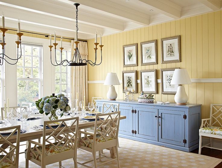 Salle à manger avec mur en bois clair jaune et plafond blanc avec deux lampadaires. Table à manger en verre avec sièges blancs sur carrelage.