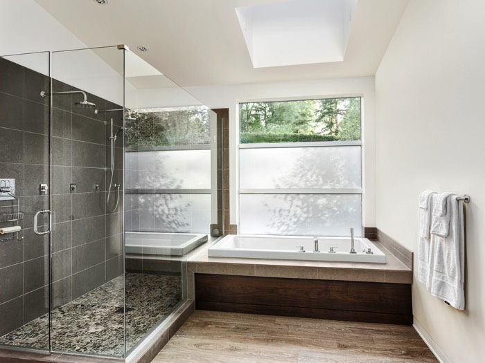 Douche à l'italienne avec deux colonnes de douche classique et un receveur surélevé en petites pierres. Baignoire blanche sur parquet en imitation noyer.