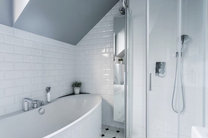 Salle d'eau avec carrelage mural tout blanc et baignoire classique. Douche avec colonne de douche classique et porte en verre transparent.